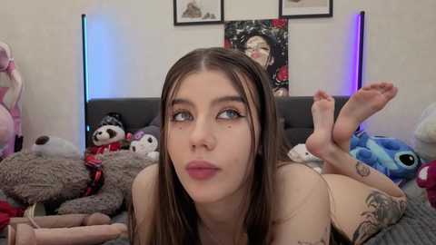 Video of a young, fair-skinned woman with long brown hair, freckles, and blue eyes, lying on a bed filled with plush toys, making a pouty face.