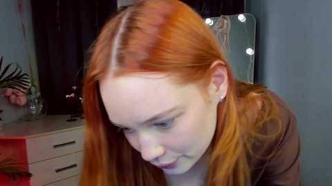 A video shows a young, fair-skinned woman with long, red hair and a brown coat, leaning forward in a softly lit, modern bedroom with a dresser, mirror, and floral arrangement in the background.