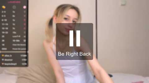 Video of a young, blonde woman with fair skin and a slim build, wearing a white tank top, seated on a bed. Background includes a calendar and beige walls.