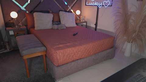 Video of a cozy, modern bedroom with a queen-sized bed covered in a quilted, peach-colored duvet, two matching pillows, and a wooden headboard. The room features a striped cushioned bench, a nightstand with a lamp, and a neon \"Lucky\" sign.