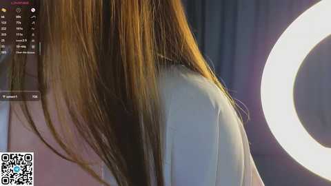 Video of a woman with long, straight brown hair, wearing a white shirt, standing in front of a large, round ring light. Background features a dimly lit room with dark curtains.