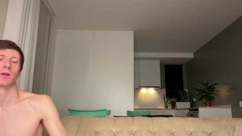 Video of a shirtless, pale-skinned, short-haired man with closed eyes, sitting on a beige tufted sofa in a modern, dimly-lit living room with white walls, a kitchen island, and potted plants.