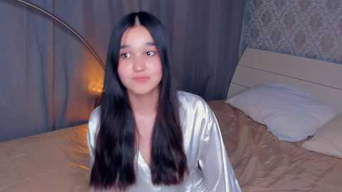 Video of a young South Asian woman with long black hair and medium skin tone, wearing a shiny silver robe, sitting on a bed with beige sheets and white pillows. The background features dark gray curtains and a patterned wallpaper.