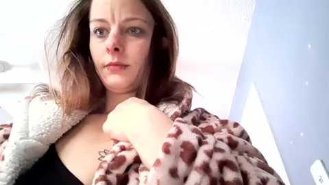Video of a young woman with light skin and shoulder-length brown hair, wearing a leopard-print robe, holding a fluffy white pillow, and looking slightly annoyed. Background shows a blue wall and a white bed frame.