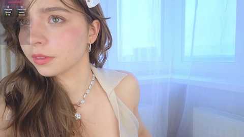 A video of a young woman with long brown hair, fair skin, and blue eyes, wearing a sleeveless top, standing indoors near a window with sheer white curtains.