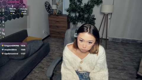 Video of a young woman with straight, shoulder-length hair, wearing a cream-colored cardigan and jeans, sitting on a gray chair in a modern living room.