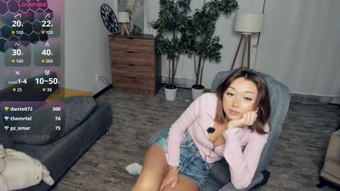 A video of an Asian woman in a pink cardigan, denim shorts, and high heels, sitting on a gray chair in a modern living room.