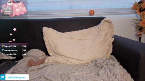 A video of a sleeping baby on a dark grey sofa, covered with a light grey blanket, with a large, fluffy pillow.