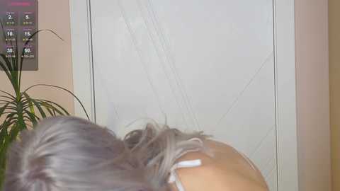 Video of a person with light gray hair, wearing a white top, standing in front of a large, slightly distorted mirror with visible cracks. The background includes a potted plant and a light-colored wall.