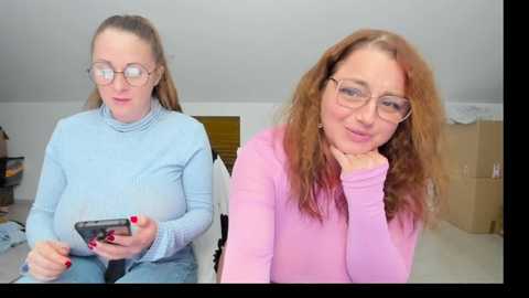 Video of two women with light skin, one with long brown hair, the other with straight blonde hair, wearing glasses and light blue and pink sweaters, seated indoors, looking at camera.