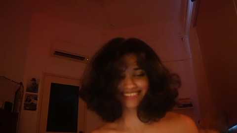 Video of a smiling woman with voluminous, curly black hair, set against a dimly lit, cluttered room with a white wall, an air conditioner, and a door.