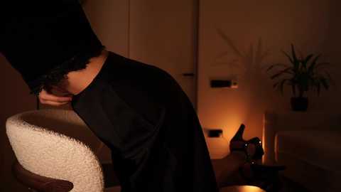 Video of a person in a black lace dress bending over a white chair, dimly lit room with a potted plant, and a candle on a wooden table.
