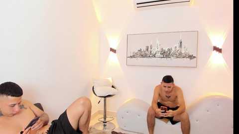 Video of a modern, minimalist room with a white wall, an air conditioner, and a black-and-white cityscape print. A shirtless man reads a book on a white couch, while another man, partially visible, lies on a nearby chair.