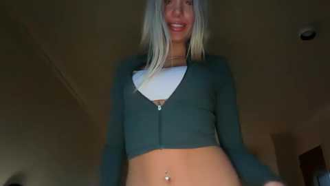 Video of a smiling young woman with long, platinum blonde hair, wearing a green cropped top and white bra, standing indoors with a plain, dimly lit background.