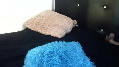 Video of a plush toy dog lying on a black bed with a fluffy blue blanket. The dog has a tan, tufted pillow and sits against a dark, tufted headboard with small, shiny buttons.
