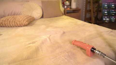Video of a beige bed with a textured duvet, a round pillow, and a red-handled screwdriver in the foreground, set in a cozy, dimly lit bedroom with a digital clock on the right.
