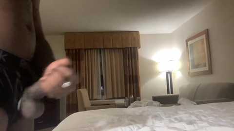 A video of a hotel room showing a blurry figure in black clothing throwing a punch, a bed with white sheets, a lamp, and beige curtains in the background.