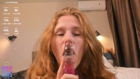 Video of a young woman with long red hair, eyes closed, using a pink dildo in her mouth. Background features a beige wall with a black light fixture, a bed, and a white air conditioner.