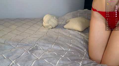 Video of a person wearing a red thong, lying on a bed with a grey quilted bedspread and a white pillow, in a dimly lit room.