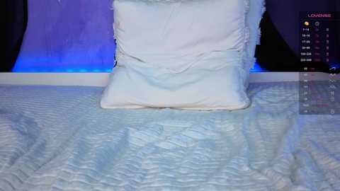 Video of a white, textured quilt with a white pillow on top, set against a blue and black backdrop with a clock displaying \"12:00.\