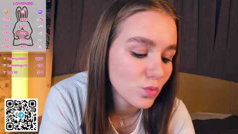 Video of a young Caucasian woman with long brown hair, wearing a light blue sweater, blowing a kiss in a cozy indoor setting with a laptop displaying a \"LoveLive!\" character in the background.