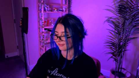 Video of a smiling, glasses-wearing woman with blue hair, wearing a black \"Wonder Woman\" t-shirt, in a dimly lit room with purple lighting, surrounded by potted plants and shelves.