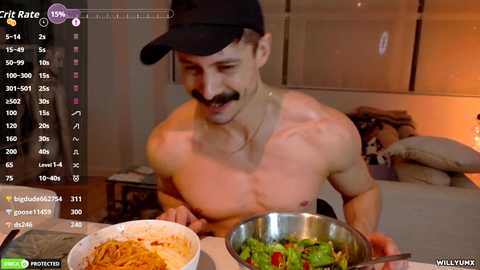 Video of a shirtless man with a mustache and black cap, cooking pasta and salad on a counter, in a dimly-lit room with a bed and window.