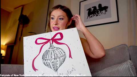 Video of a woman holding a Christmas ornament drawing with a red ribbon, in a cozy living room with a framed horse silhouette on the wall.