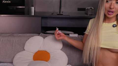 Video of a blonde woman with long hair, wearing a yellow crop top, holding a cigarette in a modern kitchen.