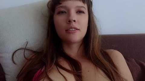 A video of a young Caucasian woman with long brown hair, light skin, and minimal makeup, lying on a brown couch against a white wall.