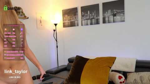 Video of a nude woman with long blonde hair, standing in a modern living room with a black leather couch, mustard and black pillows, and black-and-white photos of a cityscape on the wall.