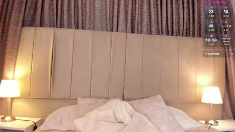 Video of a modern, minimalist bedroom with a beige, tufted headboard, two white bedside tables, and matching lamps. The room features light-colored curtains and a digital clock display in the top right corner.
