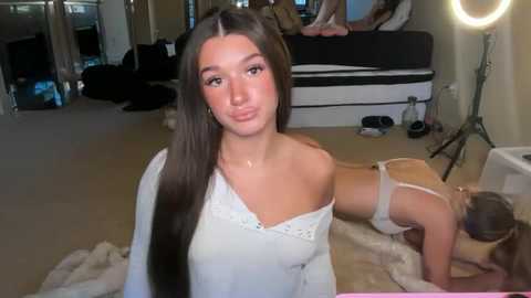 Video of a young woman with long brown hair, wearing a white off-shoulder top, standing in a dimly lit room with a plush carpet and a camera setup.