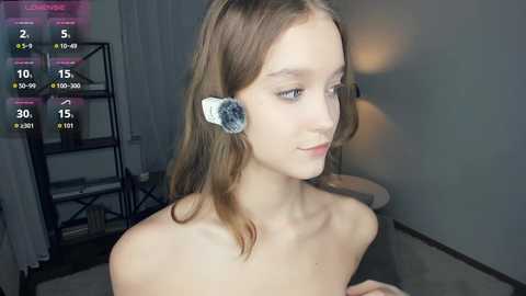 Video of a young, fair-skinned, slim woman with light brown hair, wearing a VR headset, standing in a dimly lit room with a black metal shelf and a lamp in the background.