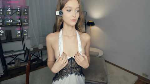 Video of a young, fair-skinned, slim woman with long, wavy light brown hair, wearing a revealing white and black lace dress with a plunging neckline, standing in a modern living room with a large flat-screen TV, shelves, and a lamp.