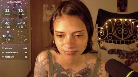 Video of a tattooed, pierced woman with dark hair, standing in a dimly lit room with a headshot camera and a metal bed.