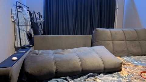 Video of a dimly lit bedroom with a beige sectional sofa, dark curtains, and a black metal clothes rack holding white shirts on the left.