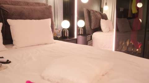 Video of a well-lit, modern bedroom with a plush, white bed, fluffy pillows, and a mirrored closet reflecting clothing. The room features a dark wooden headboard and a pair of round, glowing vanity mirrors on either side of the bed.