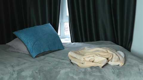 A video of a simple, modern bedroom with a gray, textured bedspread, a blue cushion, and a beige blanket folded at the foot of the bed, against dark green curtains framing a small window.