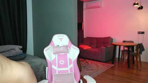 Video of a modern living room with a pink gaming chair in the foreground, a gray sofa and round dining table in the background, bathed in soft pink lighting.