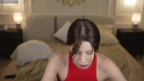 A video of a brunette woman with fair skin, wearing a red sleeveless top, leaning forward, in a dimly lit, cozy bedroom with beige walls, a bed, and lamps.