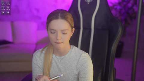 Video of a young woman with light skin and long blonde hair in a ponytail, wearing a light grey sweater, seated in a black gaming chair. Background features a purple-lit room with a beige sofa.