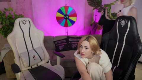 Video of a blonde woman in a white dress, leaning on a beige chair, surrounded by gaming chairs, a colorful dartboard, and a plant, in a modern, dimly-lit room.