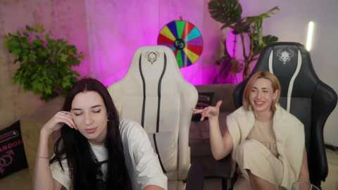 Video of two women in gaming chairs; one with long black hair, another with blonde hair, both laughing. Background includes a colorful pinwheel, green plants, and a wooden desk.