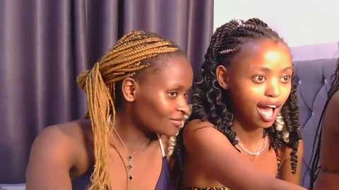Video of two African women with dark skin, one with braided hair, the other with curly hair, smiling, wearing similar sleeveless tops, against dark curtains.