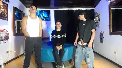 Video of three people in a dimly-lit room: two men and a woman, all wearing casual, dark clothing, standing around a pool table. The room has a mix of modern and retro decor, including posters and a drum set.