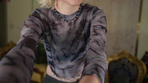 Video of a blonde woman with fair skin wearing a sheer, tie-dye crop top, black beaded necklace, and black pants, partially visible in a dimly lit, ornate room with wooden furniture.
