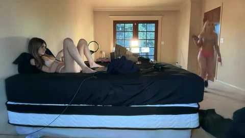 Video of three nude women on a bed, one in black lingerie, in a dimly lit bedroom with a window and a mirror.
