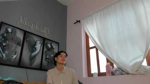Video of a young Asian man with short black hair, wearing a beige shirt, standing in a modern room with grey walls, white curtains, and a framed art piece.