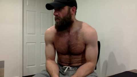 Video of a shirtless, muscular Caucasian man with a beard, wearing a black cap and gray sweatpants, sitting on a black chair in a plain, indoor setting with a white door in the background.